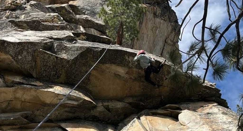 A person wearing safety gear is secured by ropes as they climb over a rock ledge high above the ground. 